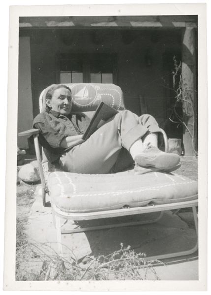 A woman, Georgia O'Keeffe, laying on a lounge chair writing a letter and smiling.