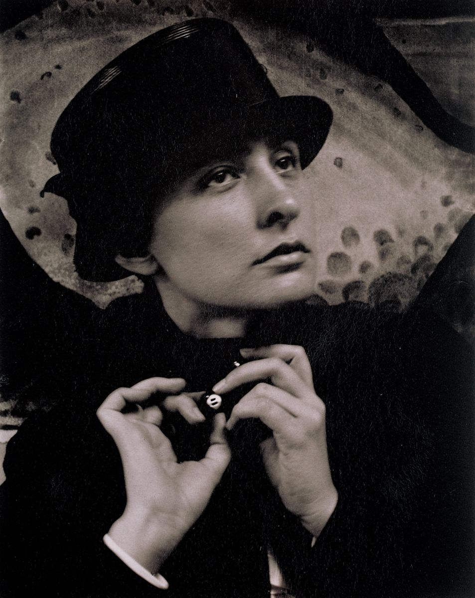 Black and white photograph. A woman, Georgia O’Keeffe, wears a dark hat and coat as she looks up and to our right, her hands gathered around the button on her high-necked collar at her throat. Her head and hands nearly fill the picture. She has dark eyes, a straight nose with a rounded end, and high cheekbones. Her mouth is closed, and her upper lip is darker than her full lower lip. The hat has a tall crown and short brim, and it comes down to her thick eyebrows and over her ears. The button of her high-collared coat shines as it catches the light. With the hand on our left, she creates an O with her forefinger and thumb as she pinches the button. Her other fingers curl in as her palm turns toward us, so her fingers resemble a cresting wave. The middle finger of that hand also touches the button, and those fingers curve toward her palm, which faces our left. Behind her is an abstract work showing a light-colored, rounded object within a darker field. The round area creates a kind of halo around her head. Her face and hands are slightly out of focus.