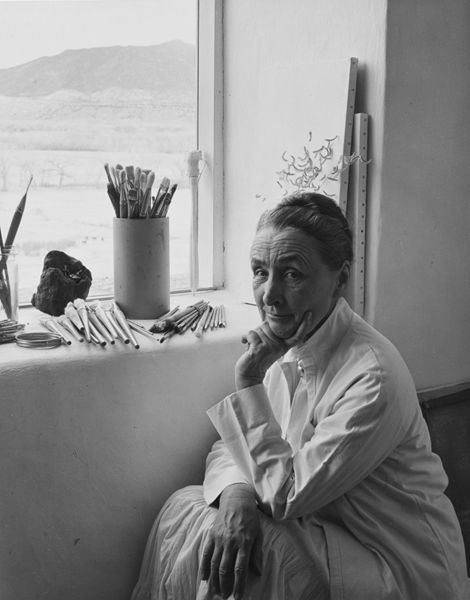 A woman, Georgia O'Keeffe, looks straight at the camera with her hand holding up her chin. She sits by a window in her artist's studio.