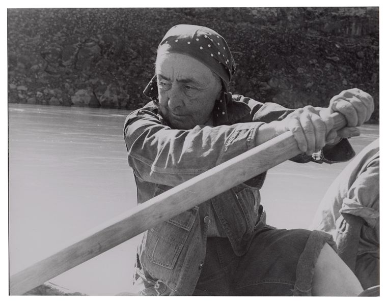 A woman, Georgia O'Keeffe, wearing a bandana on her head is rowing a boat.