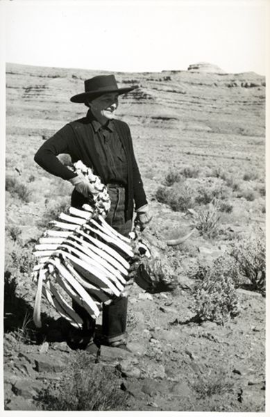 A woman, Georgia O'Keeffe, wearing all black and a black hat, holding bones of a full rib cage and partial spine while standing in the desert.